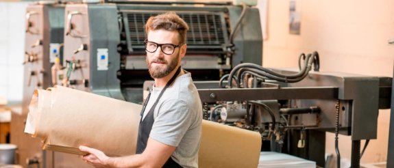 Handwerker mit Brille hält Papierrolle in Druckerei vor großer Druckmaschine