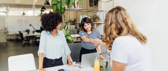 Drei Frauen schütteln sich die Hand in einem modernen Büro mit Pflanzen und Regalen im Hintergrund.