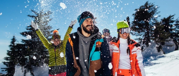 Drei Menschen werfen Schnee in die Luft und lachen in verschneiter Winterlandschaft mit Bäumen und blauem Himmel.