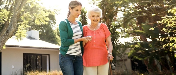 Junge Frau und ältere Seniorin gehen zusammen durch einen grünen Garten, die jüngere Person stützt die ältere liebevoll.