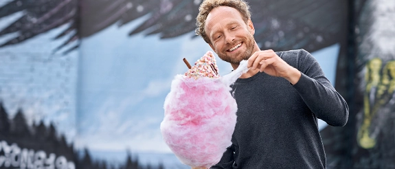 Mann mit Bart hält große rosa Zuckerwatte mit Schokolade und Streuseln vor bergiger Landschaft.