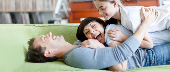 Zwei Frauen lachen zusammen auf grünem Sofa, eine sitzt hinter der anderen und umarmt sie herzlich.