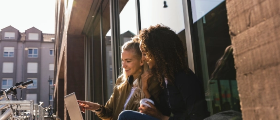 Zwei Frauen sitzen am Fenster und arbeiten gemeinsam am Laptop, Stadtkulisse im Hintergrund