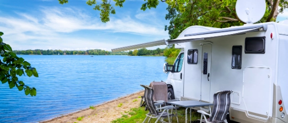 Weißer Wohnwagen am Seeufer mit Liegestühlen, Bäumen und blauem See im Hintergrund, Campingplatz.