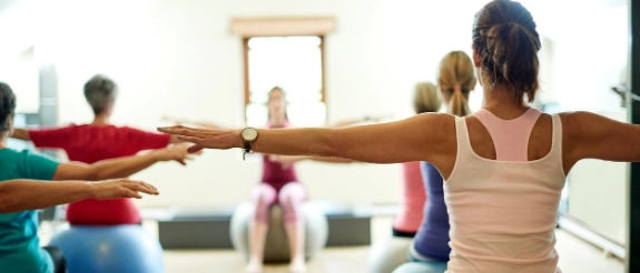 Frauen beim Pilates-Training mit Stab in heller Trainings­halle, Fitness­bälle im Hintergrund