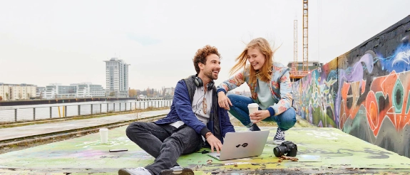 Zwei Personen sitzen am Wasser mit Laptop und Kaffee, tragen Kopfhörer, lachen zusammen mit Stadtkulisse im Hintergrund.