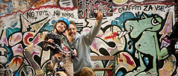 Mann und Kind auf Fahrrad vor bunter Graffiti-Wand, beide zeigen frohe Gesten
