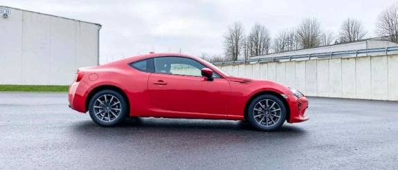 Roter Sportwagen steht auf nasser Asphaltstraße, im Hintergrund industrielle Gebäude und kahle Bäume
