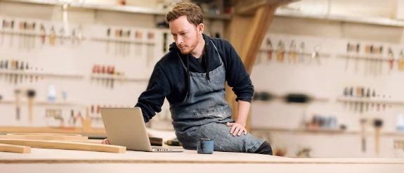 Handwerker in Arbeitskleidung sitzt an Werkbank mit Laptop, Werkstatt im Hintergrund