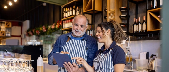 Mann und Frau in gestreiften Schürzen arbeiten zusammen in moderner Café-Küche hinter der Theke mit Regalen und Getränken im Hintergrund.