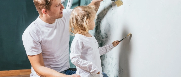 Frau und Kind streichen gemeinsam eine grüne Wand mit weißer Farbe und Pinsel.