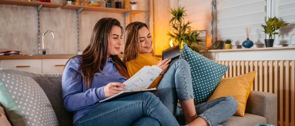 Zwei Frauen sitzen auf Sofa und schauen gemeinsam auf Tablet in gemütlichem Wohnzimmer mit Pflanzen und Kissen.