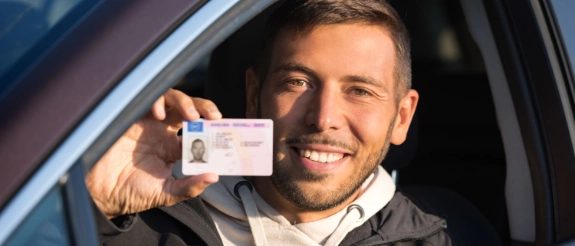 Mann im Auto hält Führerschein oder Dokument in die Kamera und lächelt.
