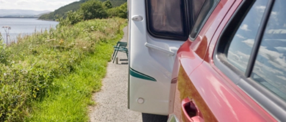 Zwei Wohnmobile parken nebeneinander auf Kiesweg, grüne Wiese und Gewässer im Hintergrund