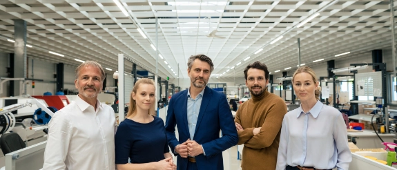 Fünf Geschäftsleute stehen sich in moderner Fabrikhalle mit Glasdecke gegenüber und schütteln Hände.