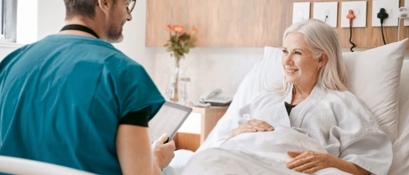 Pfleger mit Tablet spricht mit älteren Patientin im Krankenbett, beide lächeln sich an.