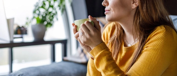 Frau mit Teetasse sitzt auf dem Sofa und schaut aus dem Fenster, sie trägt einen gelben Pullover.