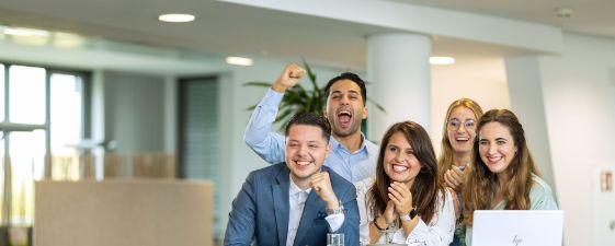 Fünf gut gelaunte Kollegen stehen in einem Büro um einen Tisch, lächeln und feiern gemeinsam. Auf dem Tisch stehen ein Laptop und ein Glas Wasser.