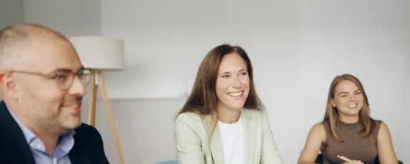 Drei Personen in Businesskleidung sitzen in einem Büro vor einem Notebook und führen ein Gespräch.