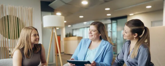 Drei Kolleginnen sitzen zusammen und unterhalten sich mit einem Tablet in der Hand.