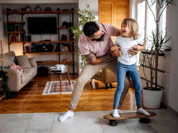 Vater bringt und junger Tochter im Wohnzimmer das Skateboardfahren bei.