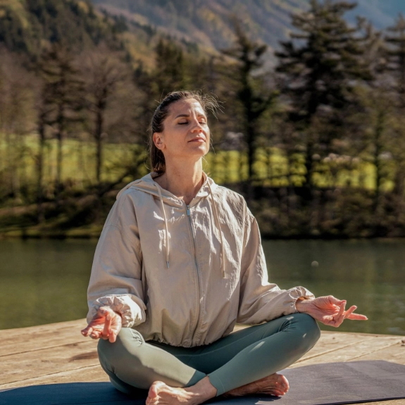 Junge Frau in Trainingsanzug sitzt auf einem Steg am See und praktiziert Yoga.
