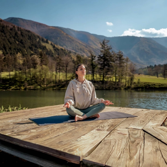 Junge Frau in Trainingsanzug sitzt auf einem Steg am See und praktiziert Yoga.