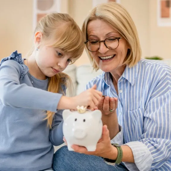 Großmutter und Enkelin werfen Münz-Geld in ein Sparschwein als Symbol für das Sparen für Kinder.