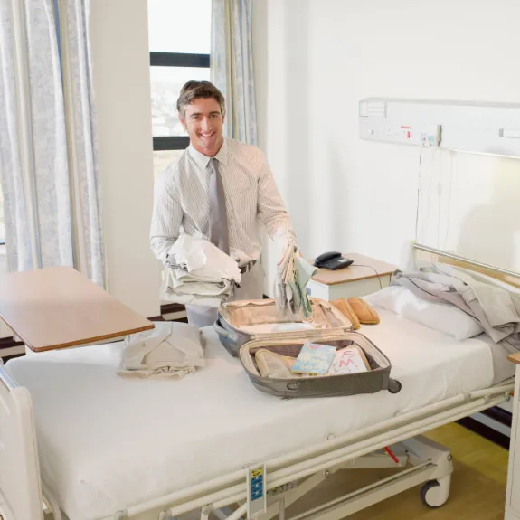 Ein junger Mann packt Kleidung in einen Koffer welcher auf einem Krankenhausbett liegt.