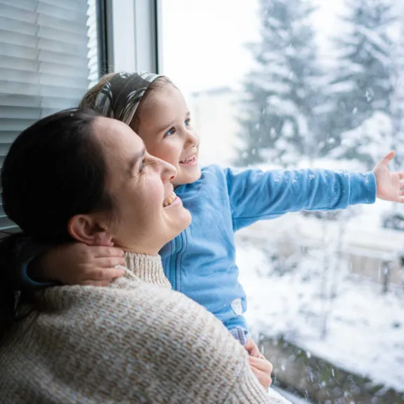 Eine Mutter hat ihr kleines Kind auf dem Arm und beide schauen aus dem Fenster. 