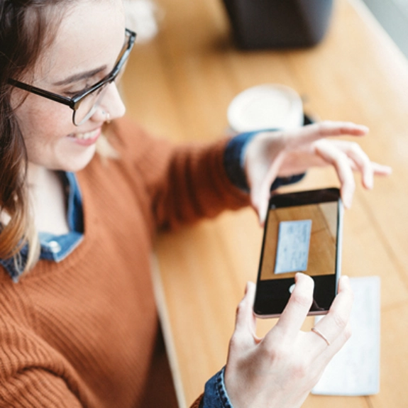 Frau mit Brille und orangefarbenem Pullover fotografiert mit Smartphone auf Holztisch