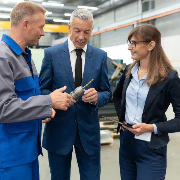 Eine Unternehmerin und ein Unternehmer stehen in einer Werkhalle und besprechen ein Projekt im Vordergrund laeuft ein Mitarbeiter an der Gruppe vorbei