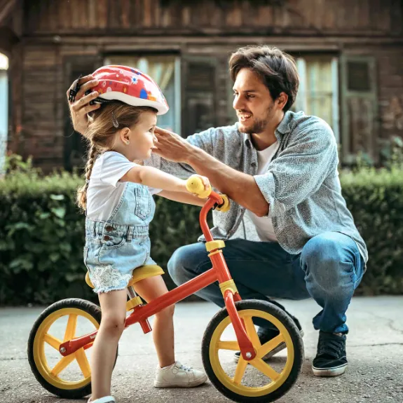 Ein Vater setzt seiner Tochter auf dem Fahrrad einen Helm auf als Symbol für die Kindervorsorge durch die Eltern. 