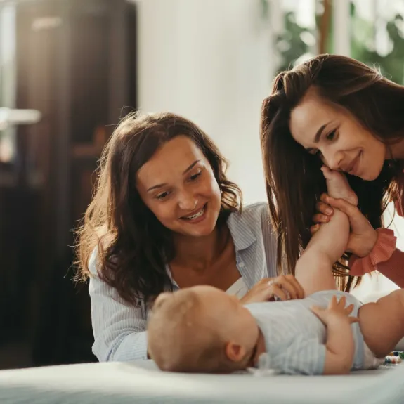 Eine junge Mutter betrachtet liebevoll gemeinsam mit einer Freundin ihr Baby. Die Freundin ist die Patentante des Babys. Sie denken über das Sparen für Patenkinder nach.