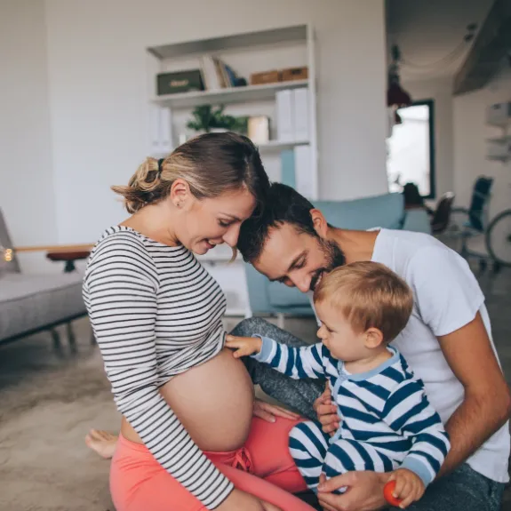 Kleiner Junge sitzt auf dem Schoss seines Vaters und fasst den schwangeren Bauch seiner Mutter an.