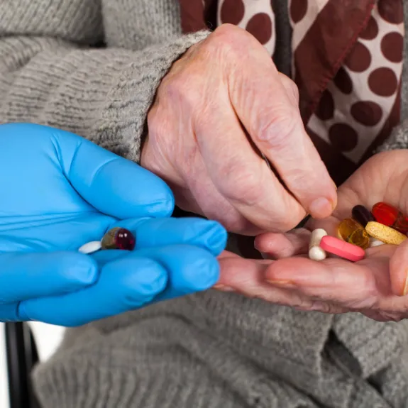 Senior mit Tabletten in der Hand nimmt noch mehr Tabletten aus der Hand einer Pflegeperson mit Handschuhen an.