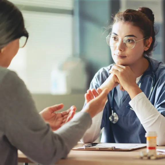 Der medizinische Dienst füllt auf einem Klemmbrett ein Formular der Pflegebegutachtung aus, während die pflegebedürftige Person mit gefalteten Händen daneben sitzt.