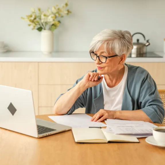 Frau sitzt am Küchentisch mit Laptop und Papieren sowie Notizbuch vor sich