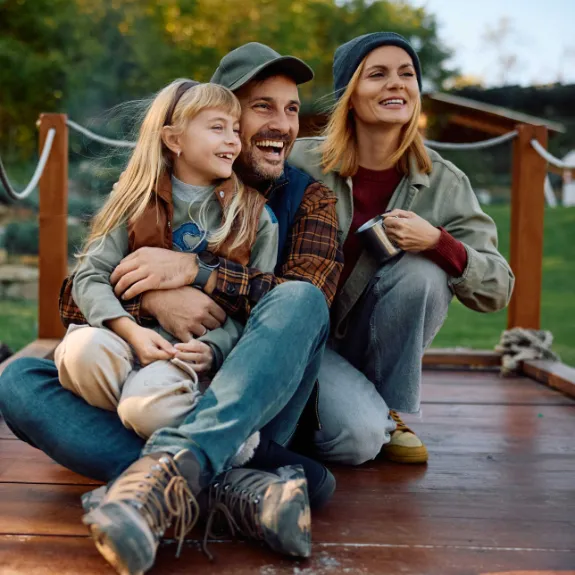 Mutter, Vater und Tochter sitzen auf einem Bootssteg und beschäftigen sich mit der Frage, was ein Kind bis zum 18. Lebensjahr kostet. 