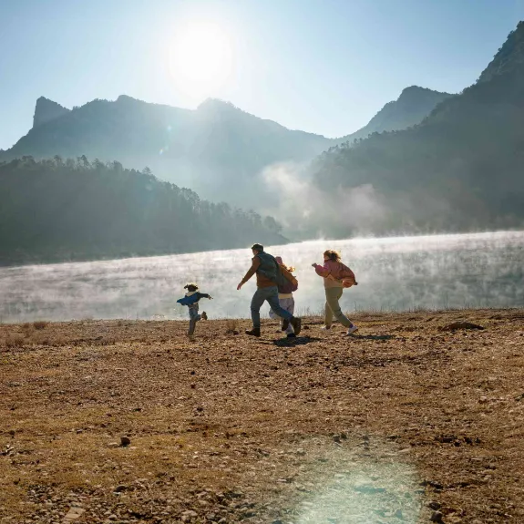 Eine junge Familie rennt fröhlich am Ufer eines Sees in den Bergen entlang.