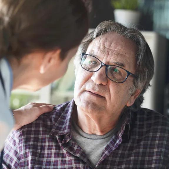 Weibliche Pflegefachperson beugt sich zu pflegebeduerftigem aelterem Mann Hand auf die Schulter legend