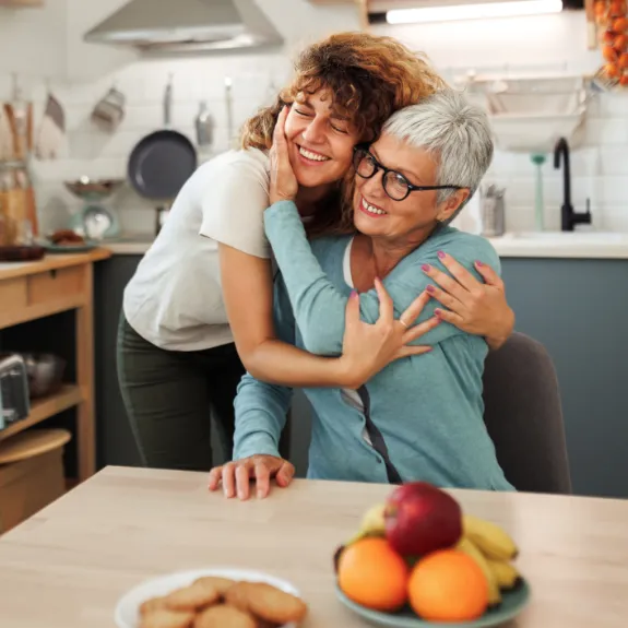 Tochter umarmt sitzende Mutter liebevoll am Esstisch.