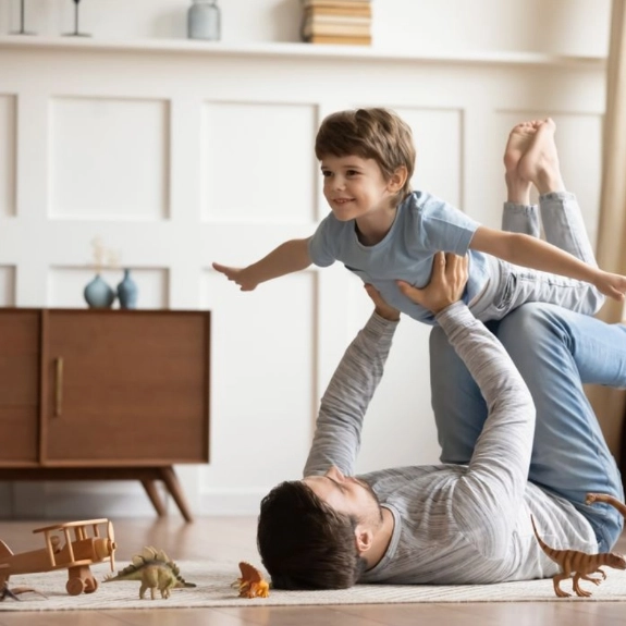 Vater liegt auf dem Rücken und hebt seinen Sohn spielerisch in die Luft im hellen Wohnzimmer
