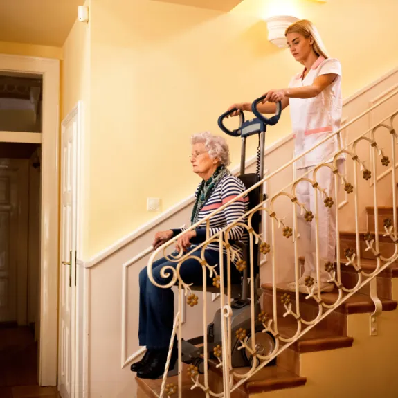Eine Pflegerin hilft einer alten Frau mit dem Sitzlift die Treppe runter.