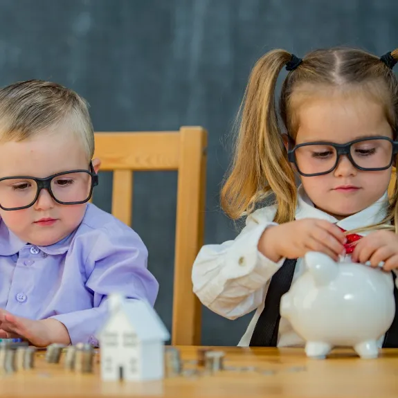 Zwei Kinder in Erwachsenenkleidung sitzen am Tisch; das Mädchen steckt Münzen in ein Sparschwein, während der Junge einen Taschenrechner hält und Münzstapel vor ihnen liegen.
