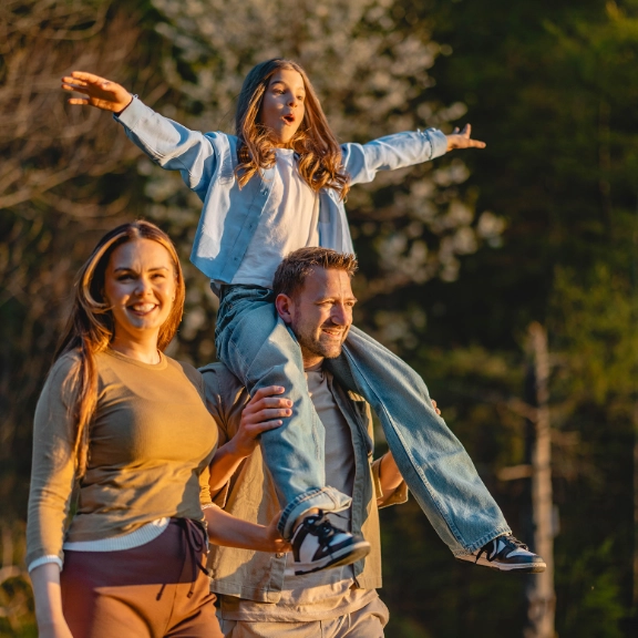 Familie wandert durch den Wald, der Vater trägt die Tochter auf den Schultern.