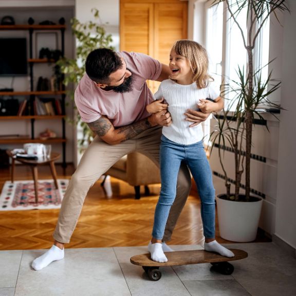 Vater bringt und junger Tochter im Wohnzimmer das Skateboardfahren bei.