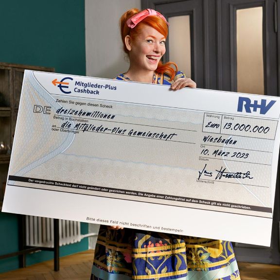 Enie van de Meiklokjes hält einen Scheck in der Hand über 13 Mio. EURO für die Mitglieder-Plus-Gemeinschaft der R+V Versicherung