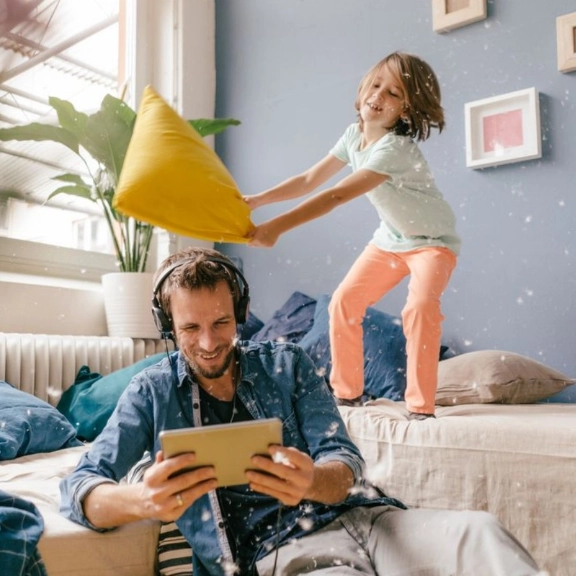 Vater hört Music auf dem Tablet und Kind tobt auf dem Sofa und spielt Kissenschlacht.
