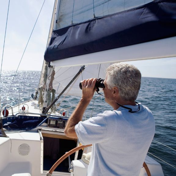 Älterer Mann steuert ein Segelboot und blickt durch ein Fernglas.